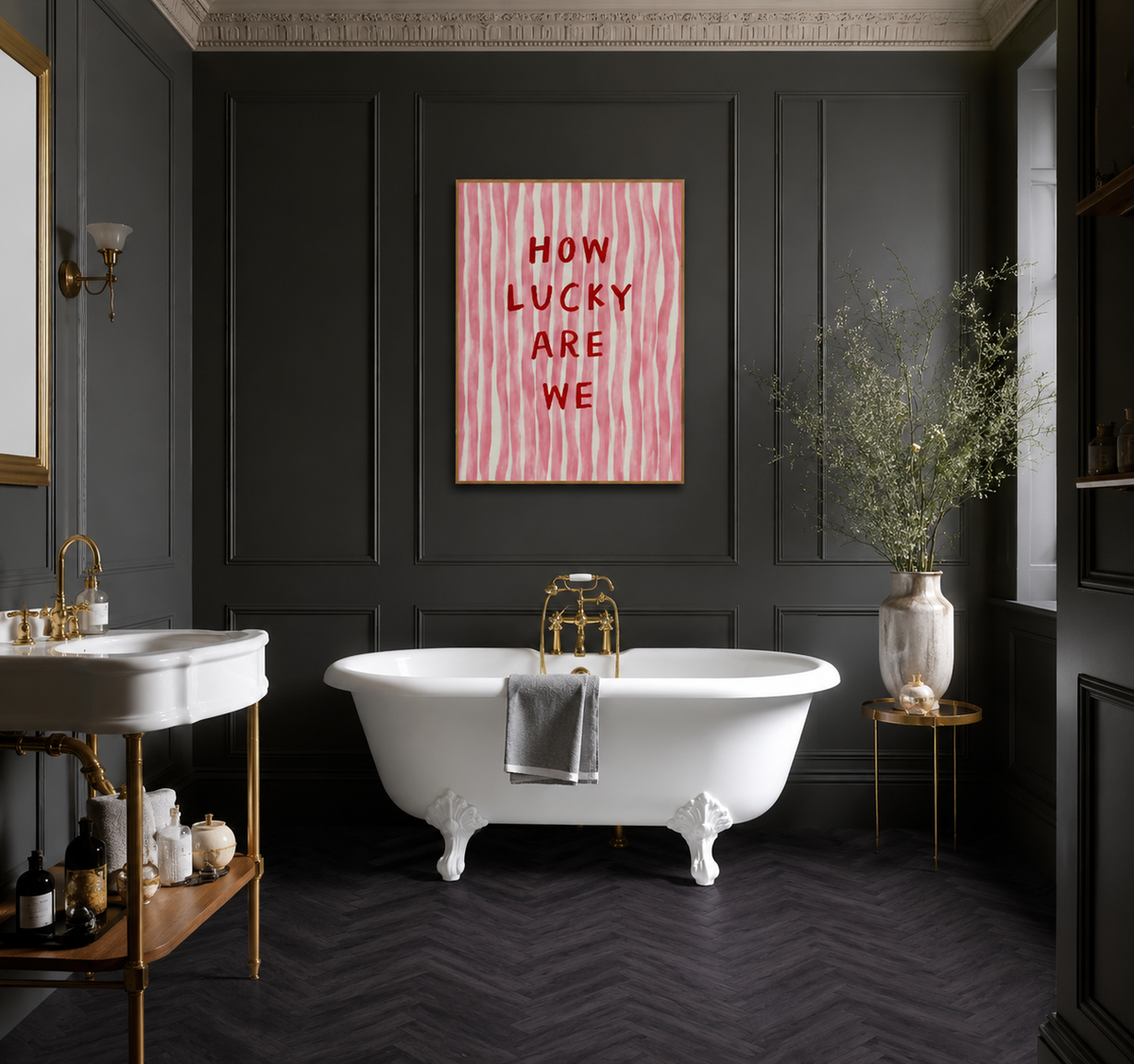Luxury bathroom featuring dark charcoal wall panelling and Black Ash herringbone flooring, with a white freestanding clawfoot bathtub, brass fixtures, pedestal sink, decorative vase with greenery, and a pink wall art print reading “How Lucky Are We.” The rich dark flooring and walls create a dramatic contrast with the bright sanitaryware and warm metallic accents.