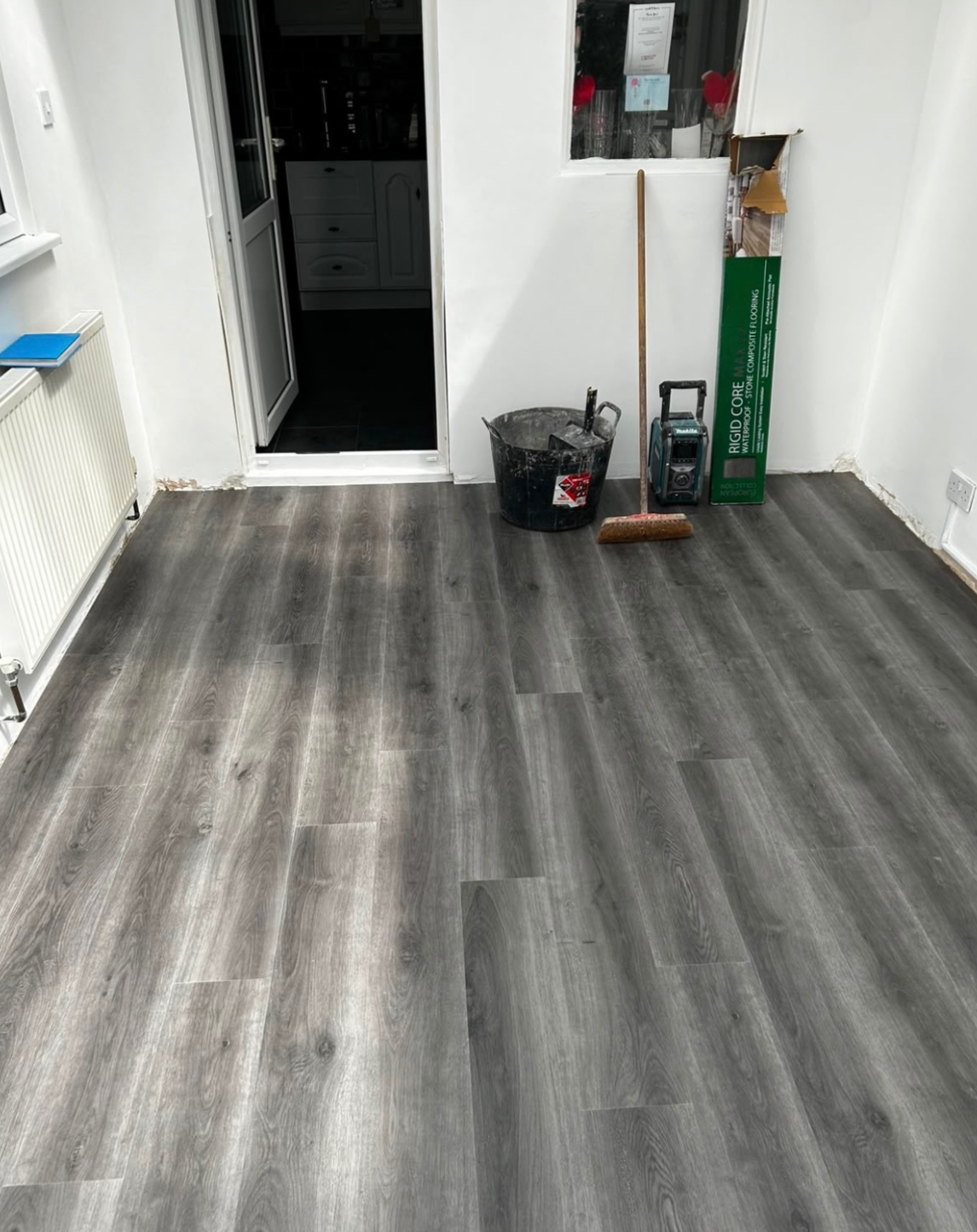 Gray wood-look vinyl plank flooring installed in a room with white walls, a doorway, and cleaning tools like a broom and dustbin in the corner.