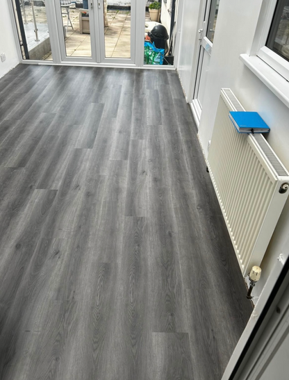 Gray wood-look vinyl plank flooring installed in a room with white walls, a white radiator, and glass patio doors in the background.