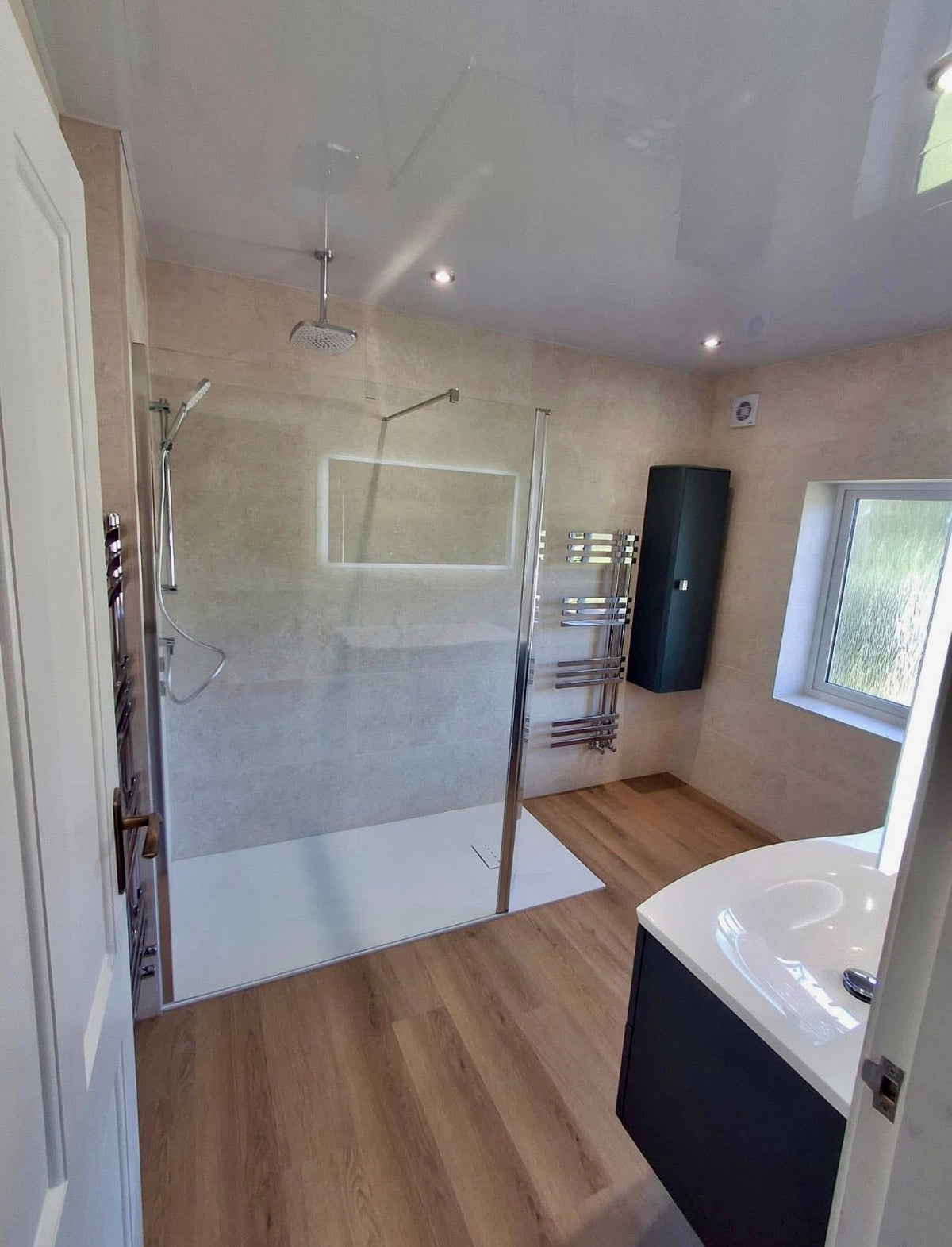 A modern bathroom interior with a walk-in shower area, wood-look flooring, beige textured walls, a white bathtub corner, and a window on the right.