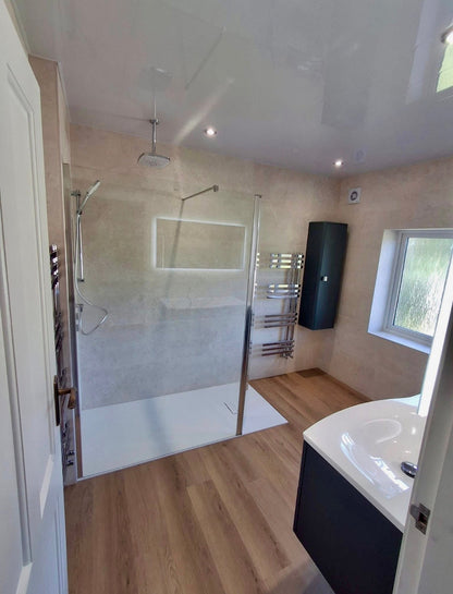 A modern bathroom interior with a walk-in shower area, wood-look flooring, beige textured walls, a white bathtub corner, and a window on the right.