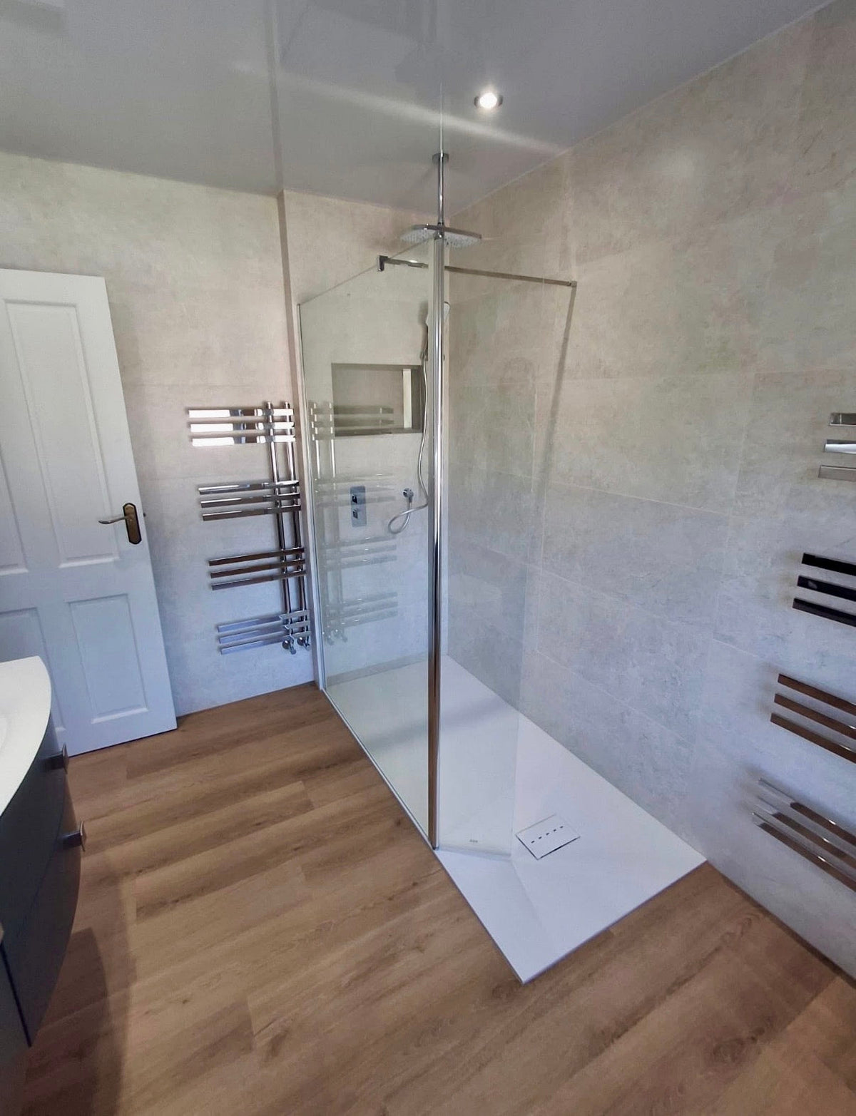 A modern walk-in shower enclosure with a clear glass door and silver metal frame, installed in a bathroom with light beige textured walls, a white shower base, and light wood flooring.