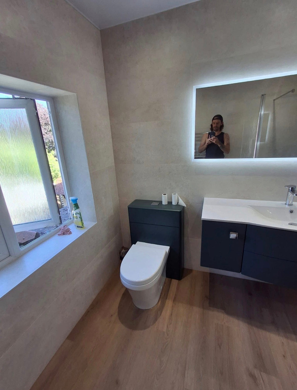 A modern bathroom interior with a white toilet, dark floating vanity with a white sink, a large backlit mirror showing a person taking the photo, wood-look flooring, and textured beige walls.