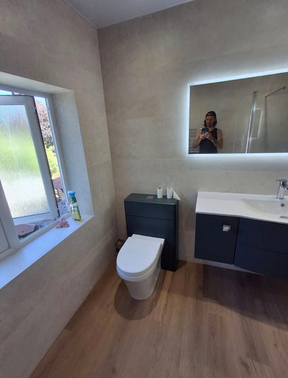 A modern bathroom interior with a white toilet, dark floating vanity with a white sink, a large backlit mirror showing a person taking the photo, wood-look flooring, and textured beige walls.