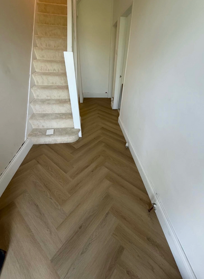 A herringbone-patterned wood floor in a hallway with white walls and a carpeted staircase on the left.