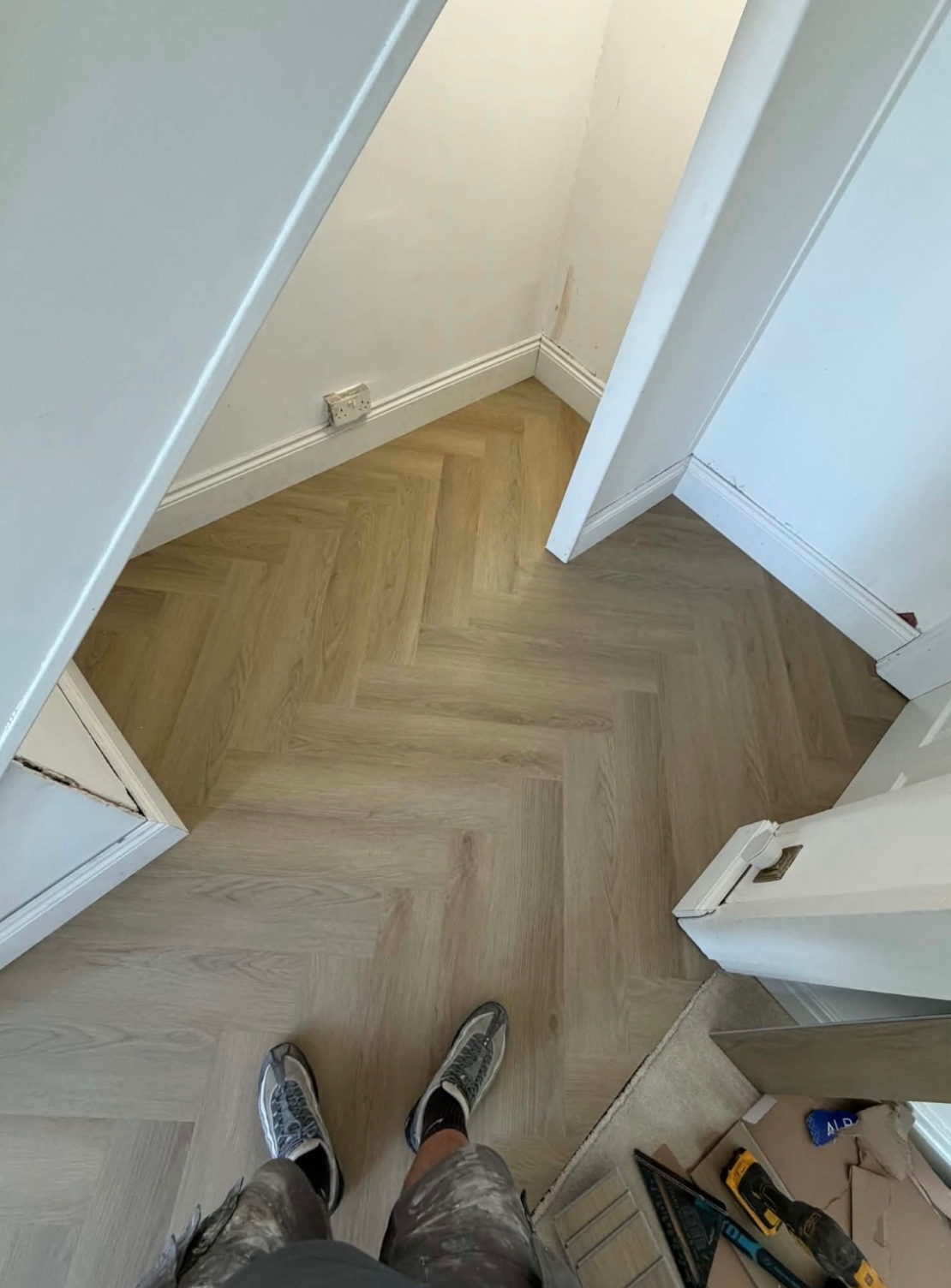 A top-down view of a person standing in a room with light oak herringbone parquet flooring, white walls, and white baseboards. The person is wearing gray slip-on shoes and gray patterned shorts.