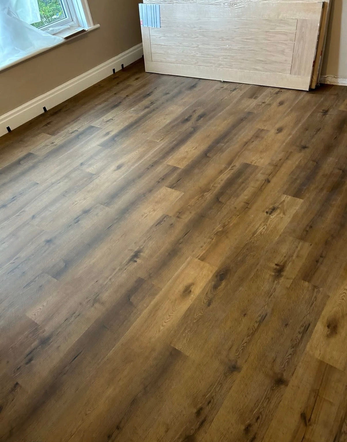 Brown wood-look laminate flooring installed in a room with beige walls, white baseboards, and a window in the upper left, with a light-colored wooden panel leaning against the wall in the background.