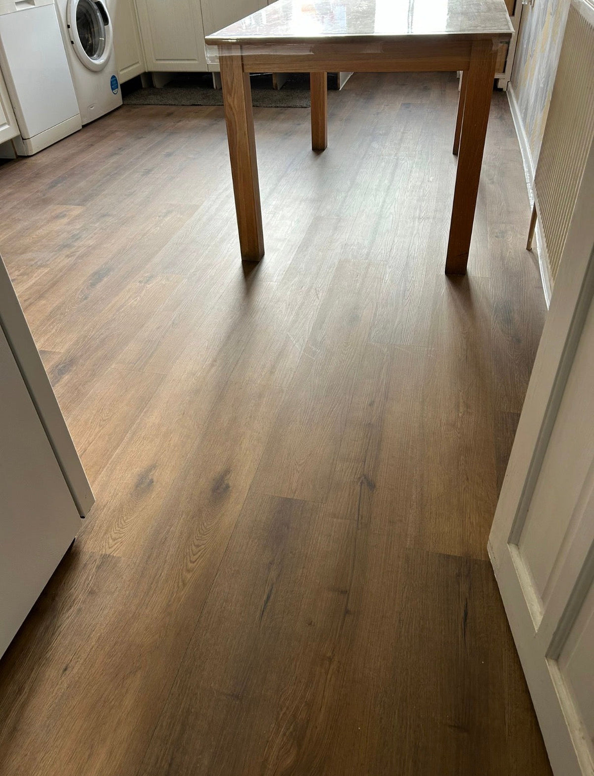 Light brown wood laminate flooring shown in a home interior setting with a wooden dining table and white appliances in the background.